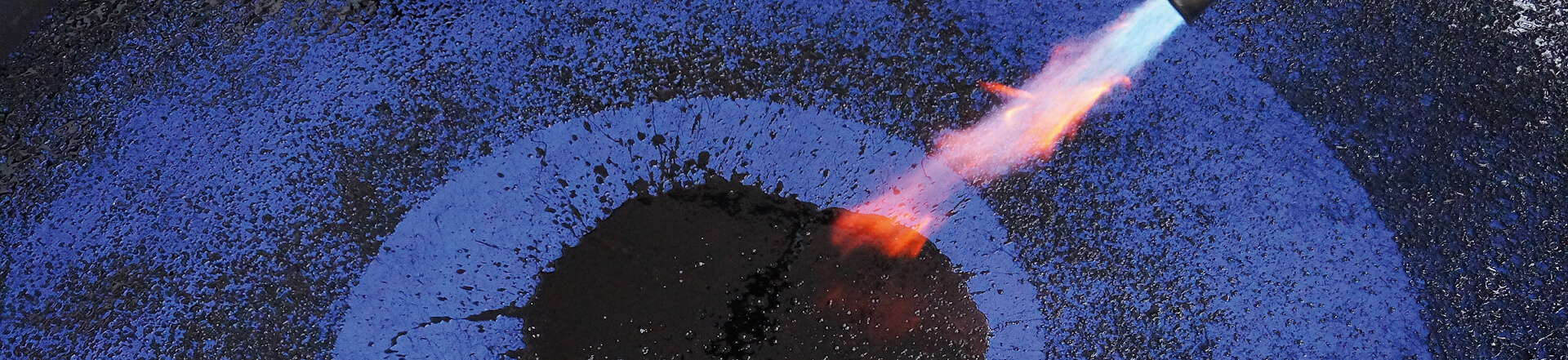 Die Flamme eines Bunsenbrenners zeigt auf ein blau-schwarzes Kunstwerk aus Asphalt von Albert Scopin